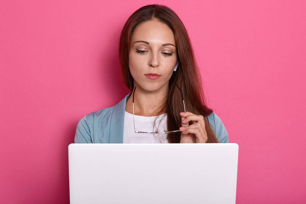 Horizontal picture of smart charming lovely young lady posing isolated over pink background in studio, holding eyeglasses in hand, working with laptop, reading text, being attentive. Study concept. - Foto, Imagem