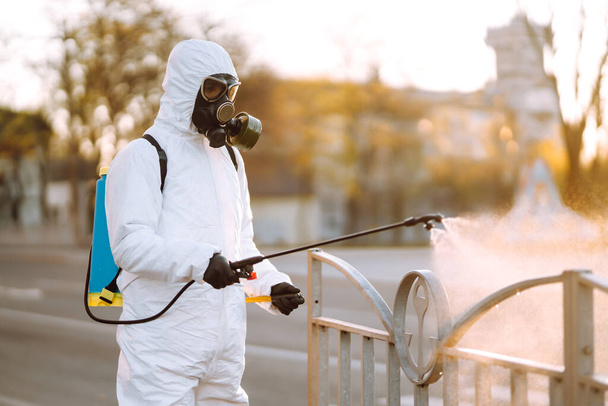 A man wearing special protective disinfection suit sprays sterilizer  in the empty public place at dawn in the city of quarantine. Covid -19. Cleaning concept. - Foto, imagen