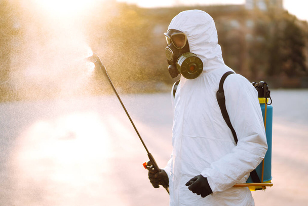 A man wearing special protective disinfection suit sprays sterilizer  in the empty public place at dawn in the city of quarantine. Covid -19. Cleaning concept. - Foto, Imagen