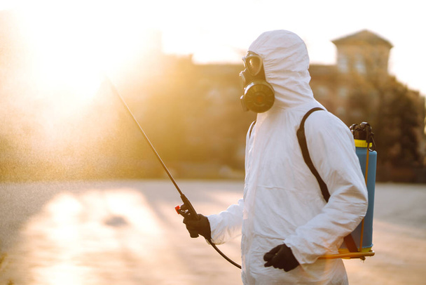 Um homem usando terno especial de desinfecção protetora pulveriza esterilizador no lugar público vazio ao amanhecer na cidade de quarentena. Covid-19. Conceito de limpeza
. - Foto, Imagem