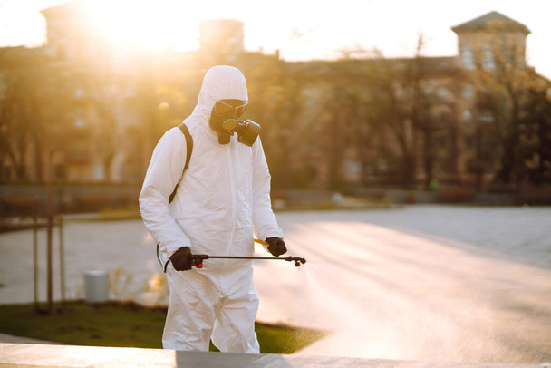A man wearing special protective disinfection suit sprays sterilizer  in the empty public place at dawn in the city of quarantine. Covid -19. Cleaning concept. - Photo, image