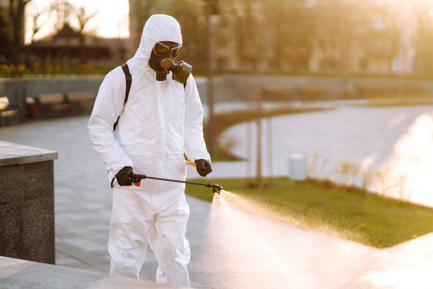 A man wearing special protective disinfection suit sprays sterilizer  in the empty public place at dawn in the city of quarantine. Covid -19. Cleaning concept. - Photo, image