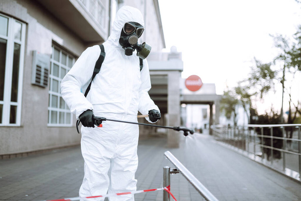 Man in protective suit  and mask sprays disinfector onto the railing in the empty public place at dawn in the city of quarantine. Covid -19. Cleaning concept. - Photo, image