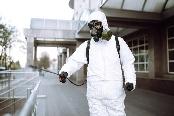 Man in protective suit  and mask sprays disinfector onto the railing in the empty public place at dawn in the city of quarantine. Covid -19. Cleaning concept. - Photo, Image