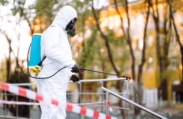 Man in protective suit  and mask sprays disinfector onto the railing in the empty public place at dawn in the city of quarantine. Covid -19. Cleaning concept. - Foto, Imagen