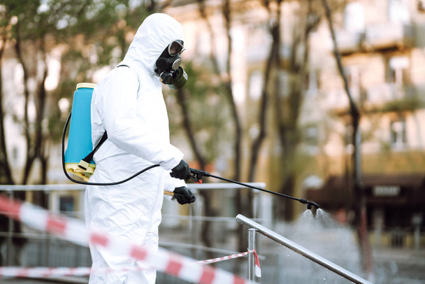 Man in protective suit  and mask sprays disinfector onto the railing in the empty public place at dawn in the city of quarantine. Covid -19. Cleaning concept. - Foto, imagen