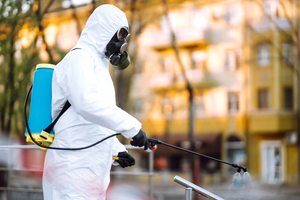 Man in protective suit  and mask sprays disinfector onto the railing in the empty public place at dawn in the city of quarantine. Covid -19. Cleaning concept. - Foto, imagen