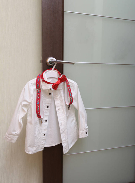 La chemise blanche intelligente d'un enfant avec une cravate et des bretelles est accrochée à la poignée de la porte
. - Photo, image