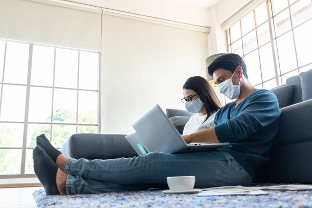 コロナウイルス発生のロックダウン中に自宅で外科用マスク作業を身に着けているカップルは、社会的距離の概念にインターネットに接続されたラップトップを使用しています - 写真・画像