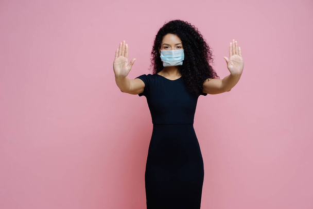 A komoly afro-amerikai nő a tenyerét a kamera felé húzza, megállítja a gesztust, elutasítja a jelet, megelőzi a koronavírust vagy a fertőző betegséget, eldobható maszkot visel, rózsaszín alapon elszigetelve - Fotó, kép