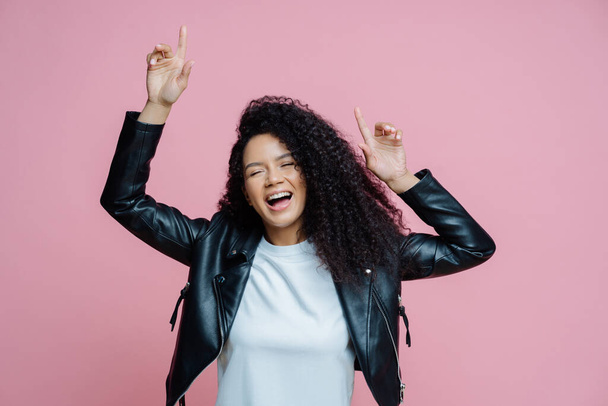 Carefree beautiful Afro American woman dances with arms raised, moves with rhythm of music on disco party, wears white t shirt and leather jacket, closes eyes with pleasure, keeps mouth opened - Foto, afbeelding