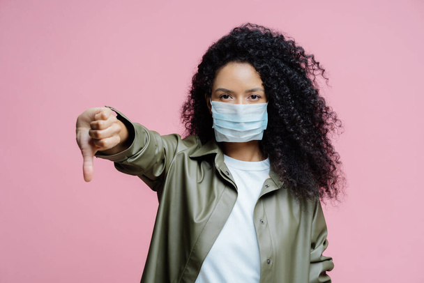 真剣な巻き毛の若い女性のイメージは、医療用マスクを身に着けている親指を下げて、ジェスチャーを嫌い、コロナウイルスの拡散を避け、健康を気にします。危険なウイルスだ。医学的概念. - 写真・画像
