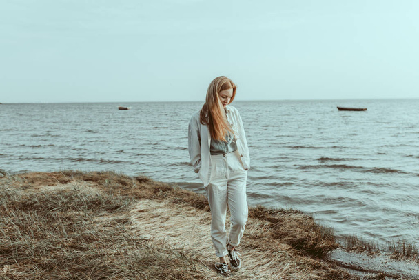 A ginger young woman walking by the lakeshore. A red headed female wearing a linen white jacket and trousers with a blue blouse. A view with water and boats. Travel concept. Slowflow fashion - Фото, изображение