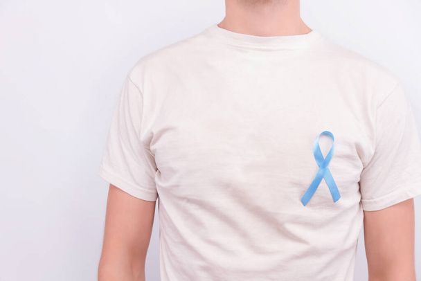 Onkolojik hastalık konsepti. Beyaz tişörtlü, açık mavi kurdeleli bir adam. Kalp kenarına prostat kanserinin sembolü.. - Fotoğraf, Görsel