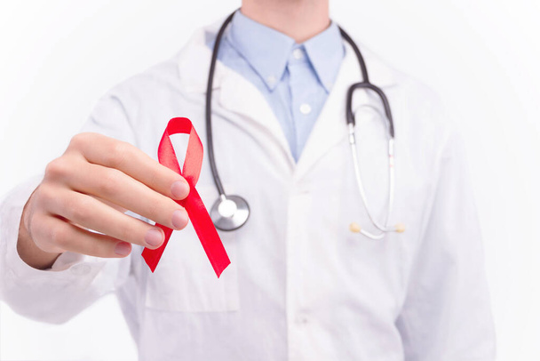 Oncological disease concept. Doctor wearing white coat and stethoscope holding red ribbon as a symbol of aids.  - 写真・画像
