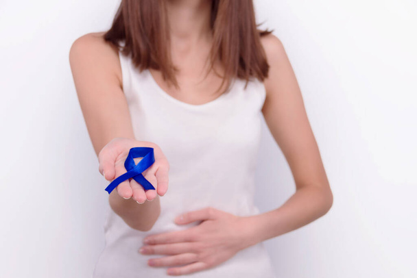 Концепція онкологічних захворювань. Дівчина носить білий верх і тримає темно-синю стрічку як символ раку кишечника і кладе руку на живіт
.  - Фото, зображення