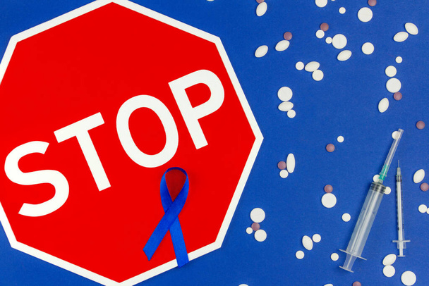 Wereldkankerdag concept. Blauw lint als symbool van darmkanker, sign stop, pillen en spuiten geïsoleerd op donkerblauwe achtergrond. - Foto, afbeelding