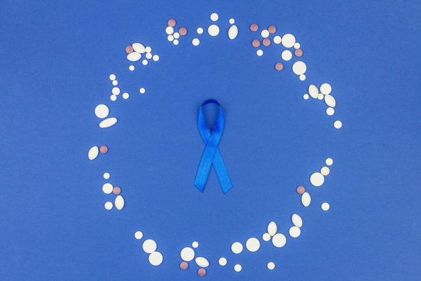 Ογκολογική έννοια της νόσου. Μπλε κορδέλα ως σύμβολο του καρκίνου του εντέρου που περιβάλλεται από χάπια που απομονώνονται σε σκούρο μπλε φόντο. - Φωτογραφία, εικόνα