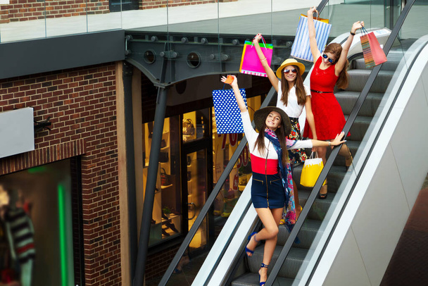 Elég boldog fényes nők női lányok barátok színes ruhák, kalapok és magassarkú bevásárló táskák séta az utcán vásárlás után bevásárlóközpontban nyáron. Bolti eladás, fekete péntek. - Fotó, kép