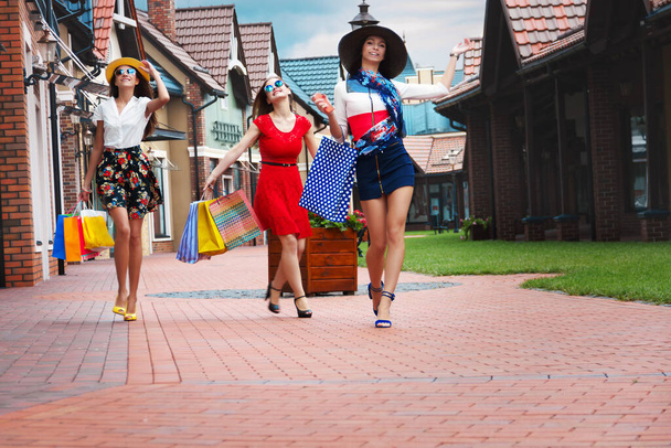 Ziemlich glückliche, helle Frauen, Freundinnen in bunten Kleidern, Hüten und High Heels mit Einkaufstaschen, die im Sommer nach dem Einkaufen in einem Einkaufszentrum auf der Straße spazieren. Ladenverkauf, schwarzer Freitag. - Foto, Bild