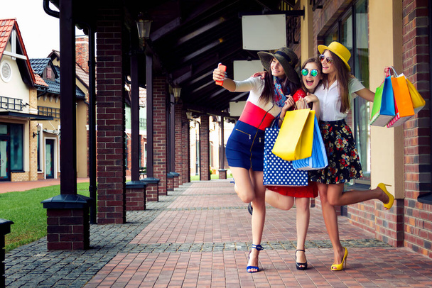 Dość szczęśliwy jasne kobiety dziewczyny przyjaciele w kolorowe sukienki, kapelusze i wysokie obcasy z torby na zakupy robi selfie po zakupach w centrum handlowym w lecie. Sprzedaż w sklepie, czarny piątek. - Zdjęcie, obraz