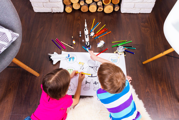 Маленькие дети рисуют костюм космонавта ручками и мечтают о космосе с помощью конструкторских игрушек космонавта: ракета, шаттл и ровер в комфортном интерьере дома на деревянном полу
 - Фото, изображение