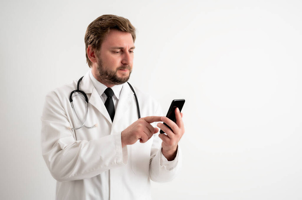 Retrato de médico varón con estetoscopio en uniforme médico con cabello castaño, sosteniendo smartphone, mensajes de texto posando sobre un fondo blanco aislado
 - Foto, imagen