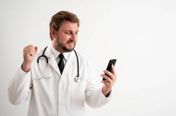Retrato de médico varón con estetoscopio en uniforme médico con cabello rubio, sosteniendo smartphone, mensajes de texto cara feliz posando sobre un fondo blanco aislado
 - Foto, imagen