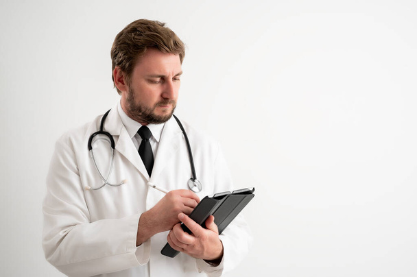 Портрет врача-мужчины со стетоскопом в медицинской форме делает заметки, позируя на белом изолированном фоне
 - Фото, изображение
