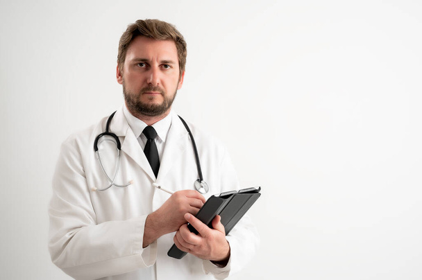 Portret pięknej kobiety lekarz ze stetoskopem w czerwonym fartuchu, z brązowymi włosami, robi notatki, patrząc na aparat fotograficzny pozujący na białym, izolowanym tle - Zdjęcie, obraz