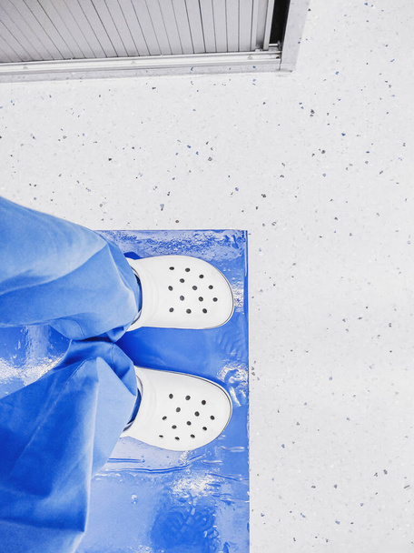 Top view of the legs of a medical worker in white slippers and blue trousers at the entrance to the laboratory. - Φωτογραφία, εικόνα