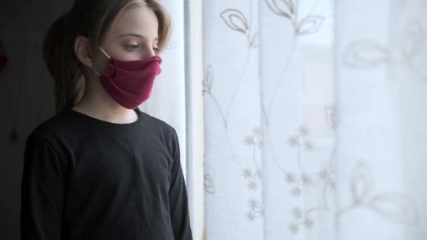 Video of sad girl wearing a cloth face mask looking through window during worldwide virus epidemic. - Video, Çekim