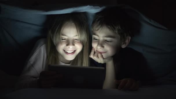 Video of cute small boy and girl lying under the blanket at night and watching video on tablet computer. - Záběry, video
