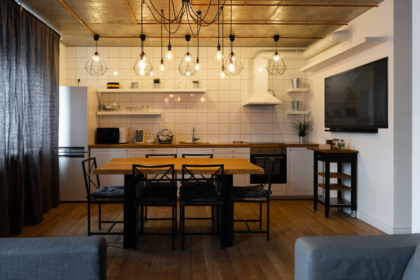 Εσωτερική διακόσμηση κουζίνας σε μοντέρνο σκανδιναβικό στυλ με μεγάλο ξύλινο τραπέζι και καρέκλες στο πάτωμα από ανοιχτόχρωμο ξύλο, λευκούς τοίχους και έπιπλα με τηλεόραση, συσκευές και κρεμαστά φωτιστικά - Φωτογραφία, εικόνα