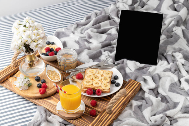 Деревянный поднос со стаканом апельсинового сока и сэндвичем с ягодами на кровати. Работа из дома утром со здоровым домашним завтраком
 - Фото, изображение
