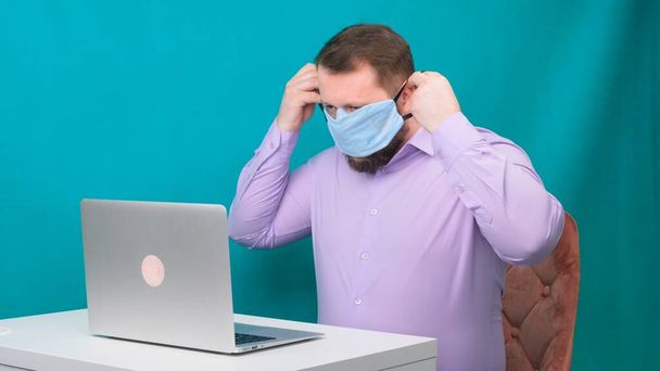 Бородатий чоловік у фіолетовій сорочці носить захисну медичну маску для роботи в ноутбуці вдома на зеленому тлі. Концепція карантину
 - Фото, зображення