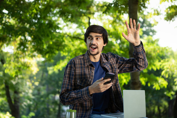 Mladý brunet na volné noze s vousy a knírkem sedí s laptopem v parku a zvedá ruku v gestu pozdravu. Práce na jakémkoliv místě - Fotografie, Obrázek