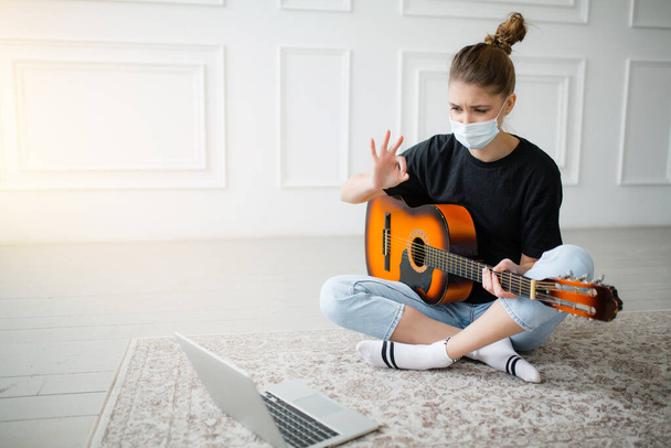 A quarantined masked girl practices guitar lessons on a laptop through a video conference with a teacher in a bright room. - Foto, imagen