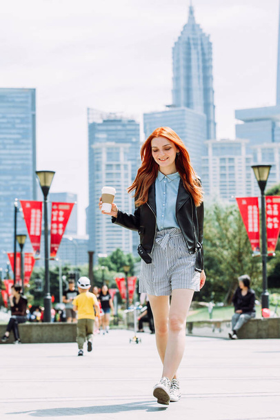 Happy young trendy red hair woman wearing black leather jacket and shorts, drinking take away coffee and walking in an urban city. City walk lifestyle concept. - Foto, Imagem