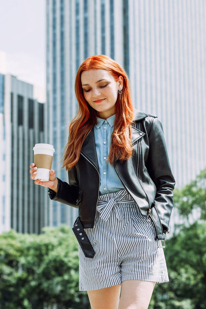 Happy young trendy red hair woman wearing black leather jacket and shorts, drinking take away coffee and walking in an urban city. City walk lifestyle concept. - Photo, Image