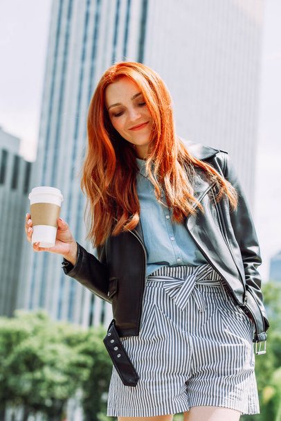 Happy young trendy red hair woman wearing black leather jacket and shorts, drinking take away coffee and walking in an urban city. City walk lifestyle concept. - Фото, изображение