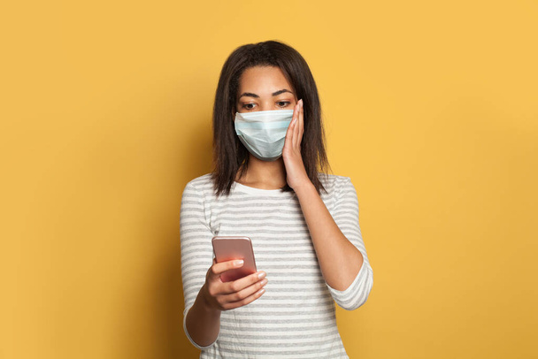Schockierte schwarze Frau mit medizinischer Gesichtsmaske erhielt Nachricht auf buntem gelben Hintergrund - Foto, Bild