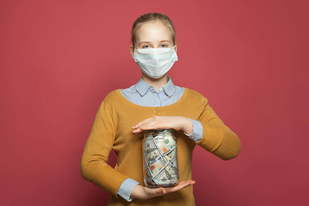 Девушка в медицинской маске держит деньги наличными нам доллары на синем фоне
 - Фото, изображение