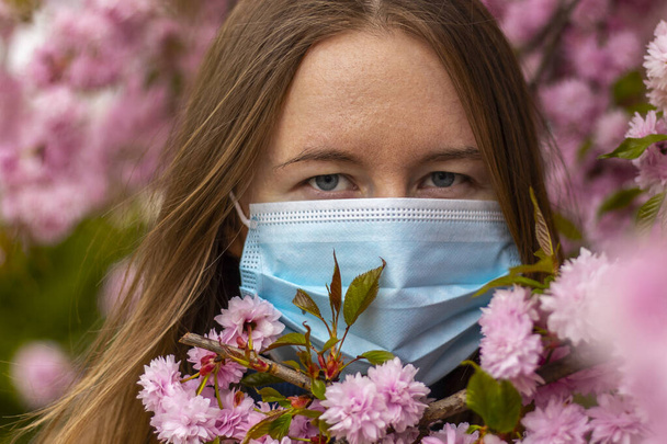 Κοντινό πλάνο πορτρέτο της όμορφης θλιμμένης γυναίκας με την ιατρική μάσκα ανάμεσα σε ροζ λουλούδια σακούρα. Φυσικό φως ημέρας. - Φωτογραφία, εικόνα