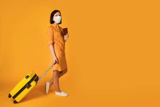 Frau mit medizinischer Gesichtsmaske hält Koffer auf leuchtend gelbem Hintergrund - Foto, Bild
