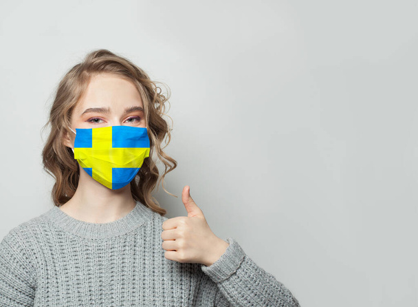 Gelukkige vrouw in het gezicht masker houden duim omhoog met Zweden vlag achtergrond. Begrip griepepidemie en virusbescherming - Foto, afbeelding
