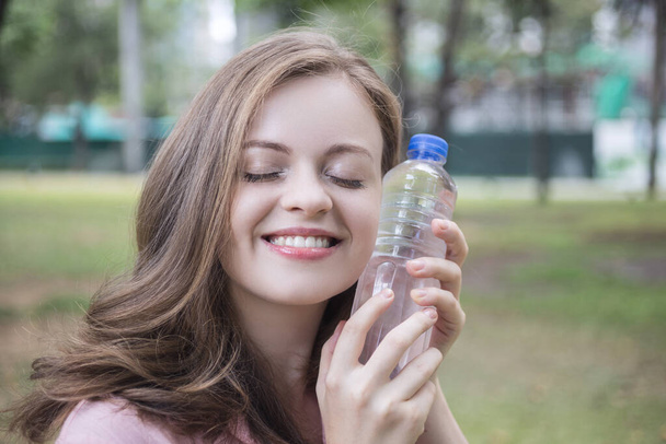 Улыбающаяся юная кавказская девушка, держащая питьевую воду из пластиковой бутылки в зеленом парке
 - Фото, изображение