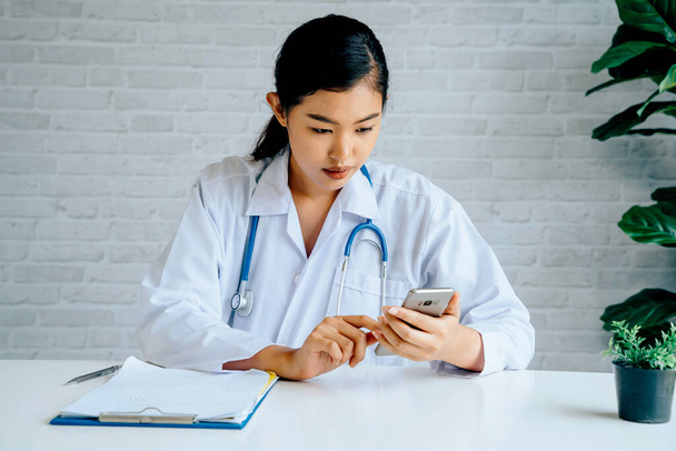 Young Asian female Doctor using a mobile phone at hospital. Healthcare worker scrolling and typing text messages at office - Fotografie, Obrázek