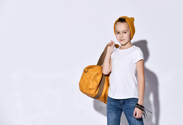 Teenage kid with orange travel bag, in hat, t-shirt, blue jeans, bracelet. Smiling, showing victory hand, posing isolated on white - Foto, Bild