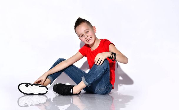 Teenage kid in red t-shirt, blue jeans, black sneakers. Sitting on floor, smiling, showing victory signs, posing isolated on white - Fotografie, Obrázek